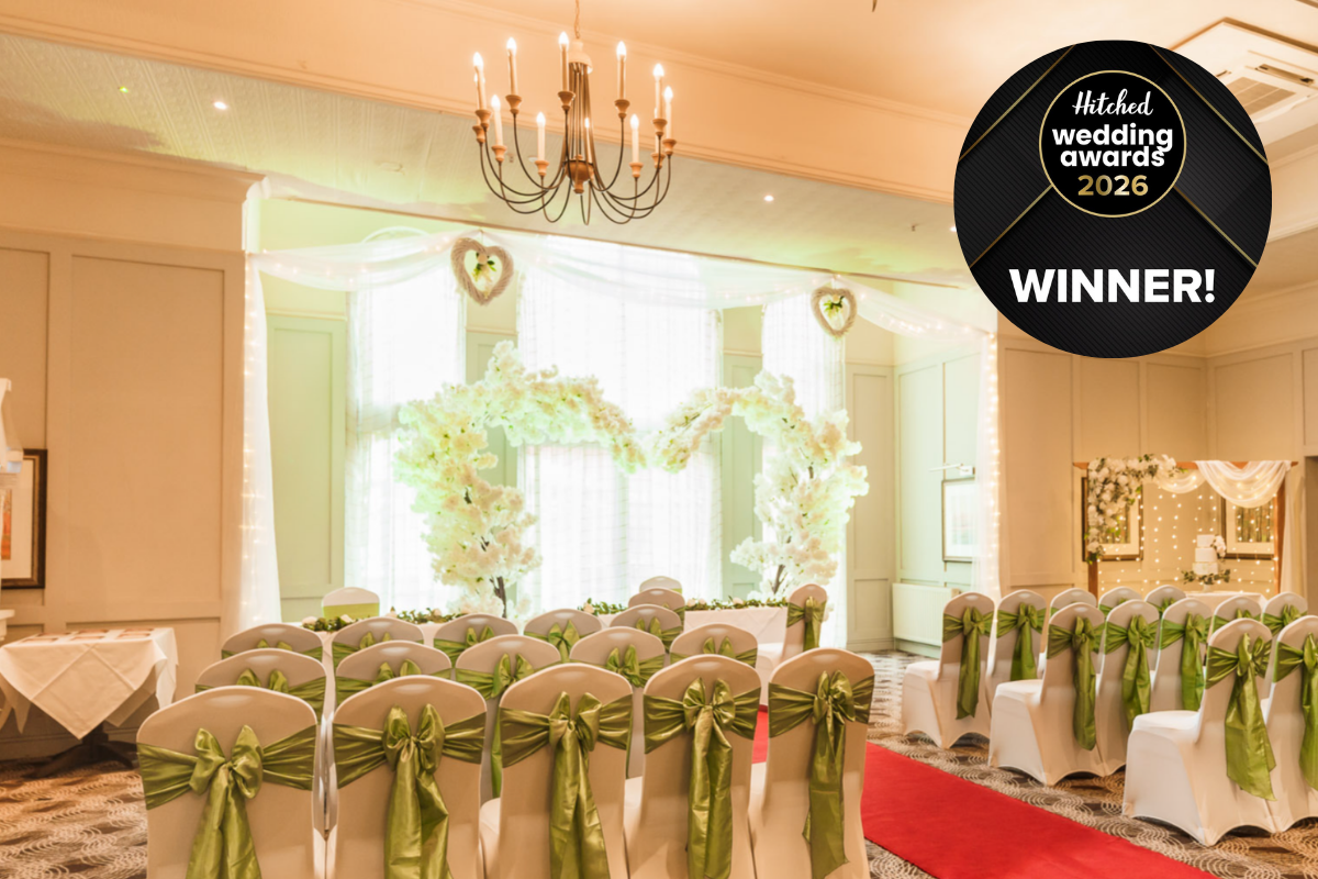A large room set up for a wedding ceremony. At the front of the room is a heart shaped arch of white flowers. There are dozens of chairs covered in white cloth and tied with green ribbons.